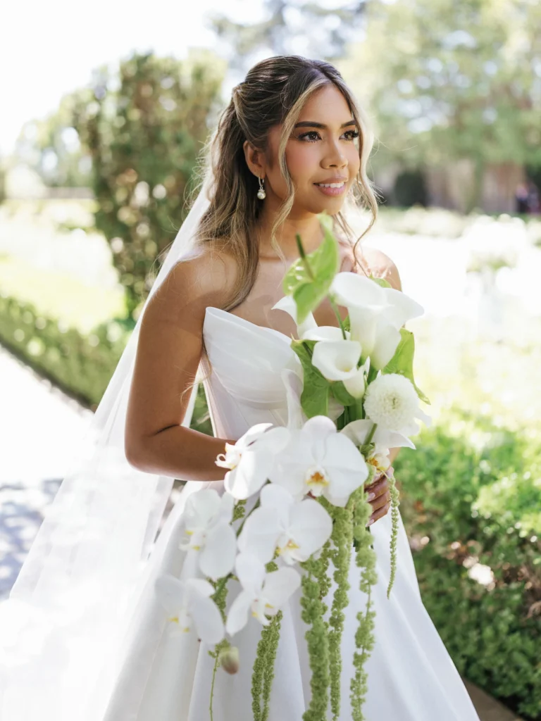 Vertical close-up portrait of a bride looking away, showcasing her white corset bodice dress and drop earrings against a blurred green hedge backdrop.
