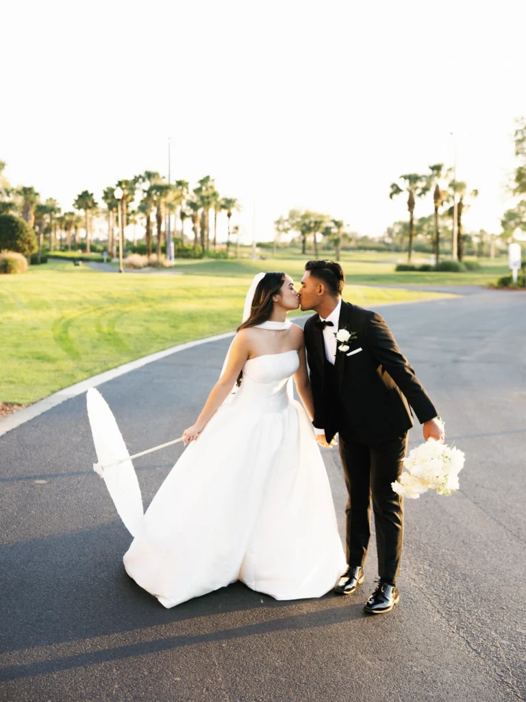 Newlyweds kissing on a paved country club path lined with palm trees, featuring a bride holding a white lace parasol and a groom in a black tuxedo.