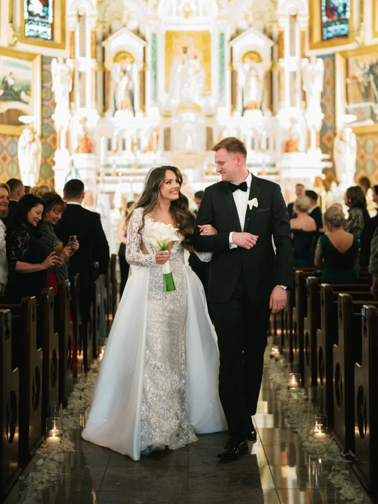 Bride in a fitted lace wedding gown with long sleeves and a detachable satin overskirt holding hands and walking down the aisle with a groom in a black tuxedo inside the ornate interior of St. John Cantius Church.