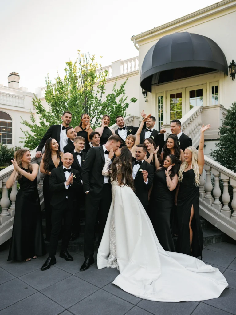 A kissing bride and groom surrounded by a large bridal party in black gowns and tuxedos cheering, laughing, and throwing hands in the air for a fun photo and film moment.