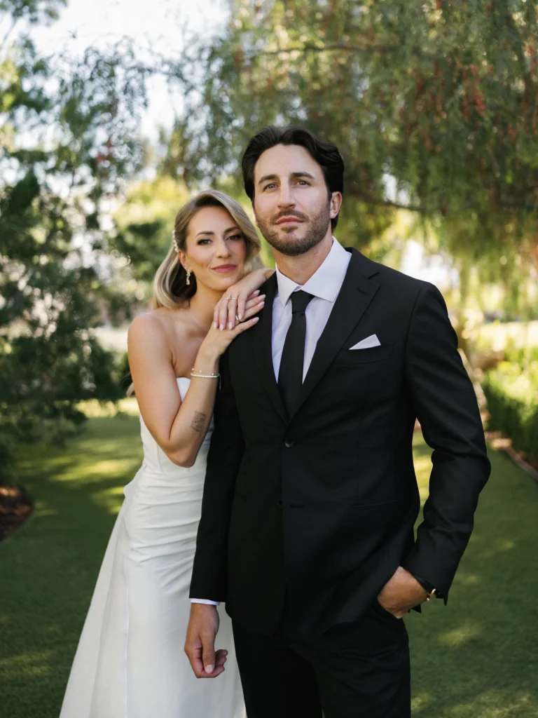 Elegant bride in a strapless silk wedding dress leaning on her groom in a tailored black tuxedo for an outdoor editorial portrait.