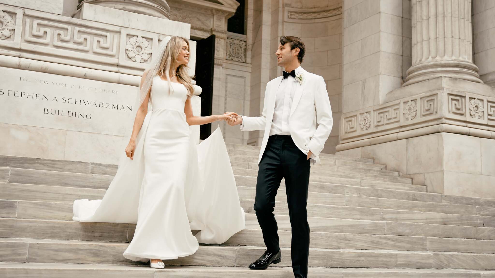 Couple holding hands on wide pale stone steps outside an architectural building.