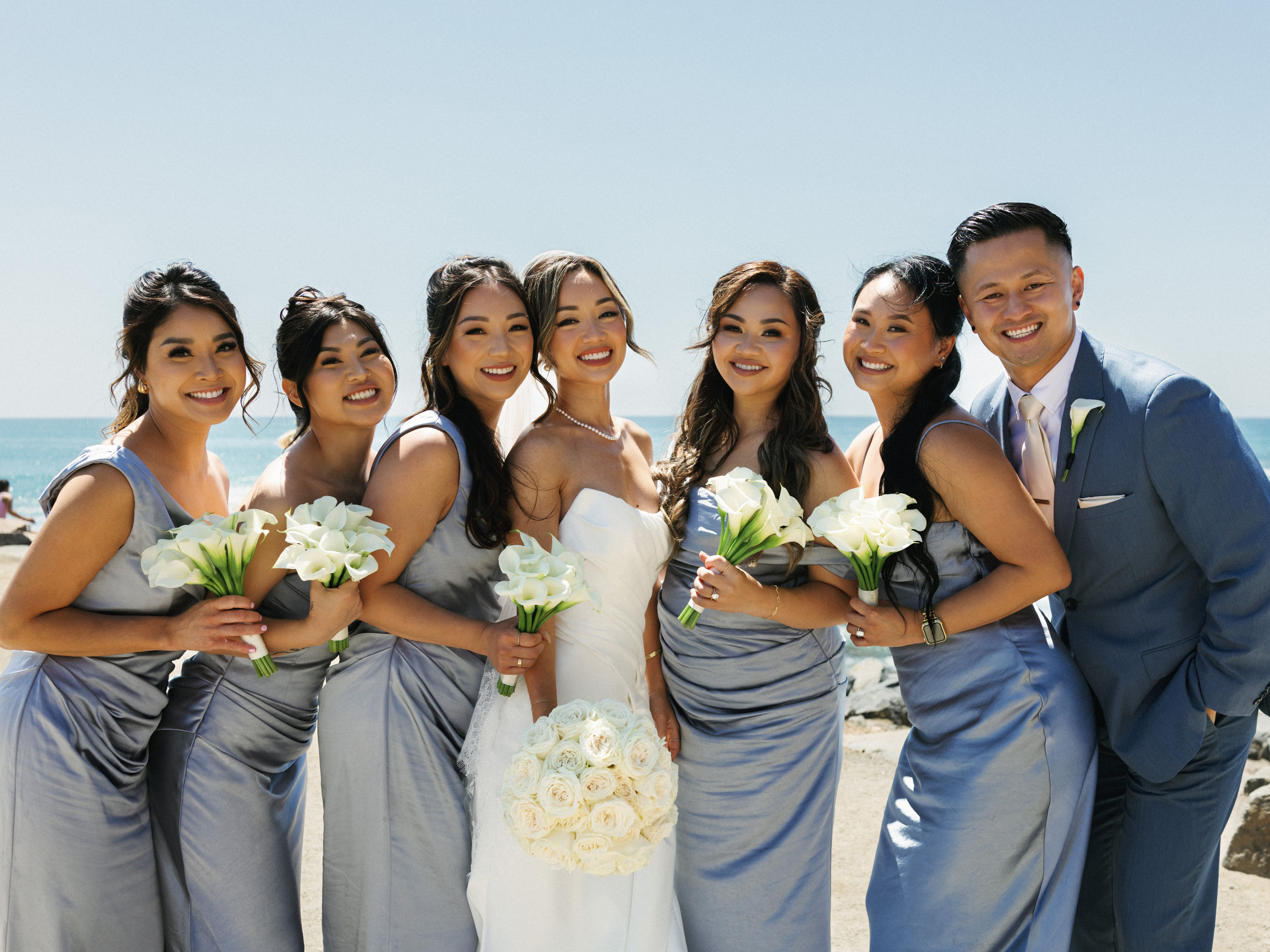Smiling bridesmaids in dusty blue silk dresses standing with the bride on a sunny beach