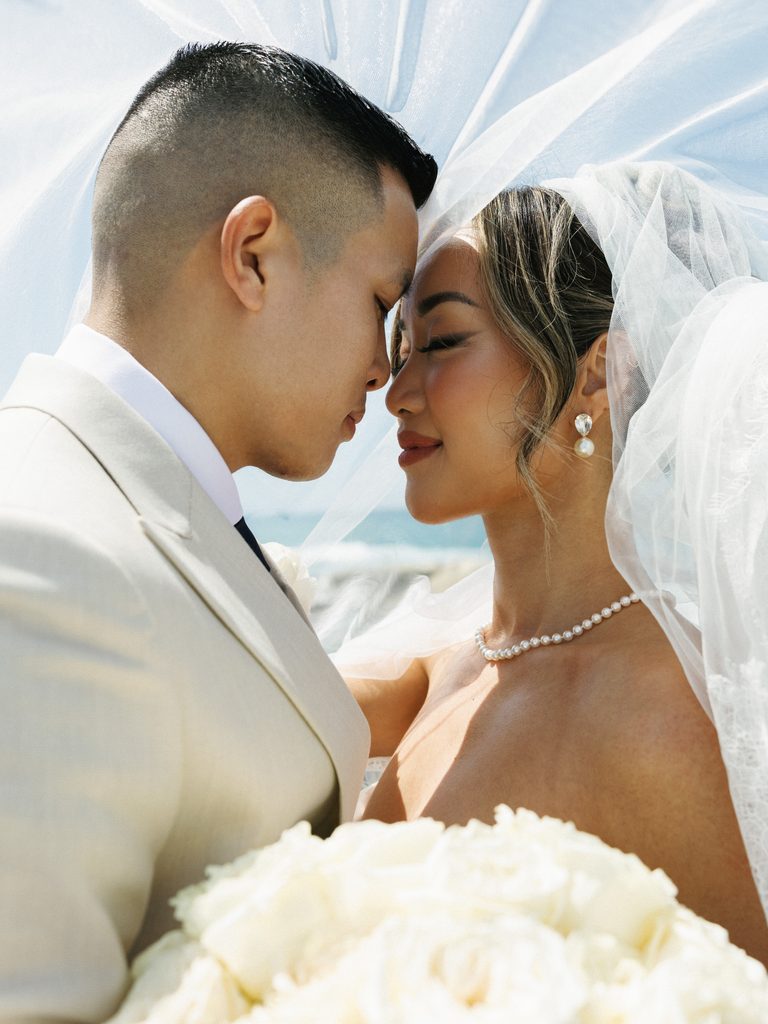 Romantic beachfront couple portrait under a flowing veil, showcasing light and airy wedding photography styles.