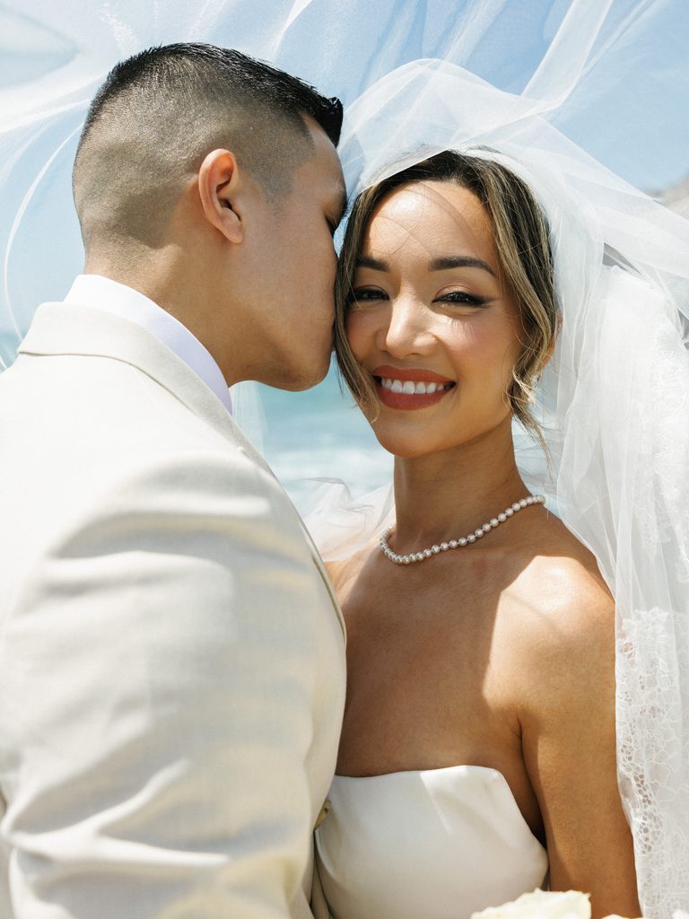Happy newlyweds sharing a candid moment outdoors, highlighting the bride's elegant pearl necklace and classic veil styling.