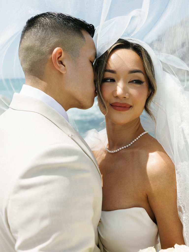 Cinematic shot of a groom kissing his bride against a soft ocean background with a blowing tulle veil.