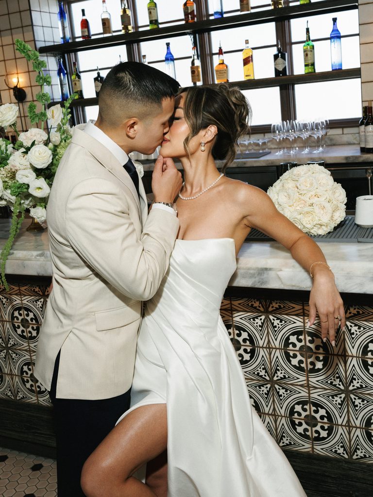 Fashion-forward couple kissing at a marble bar counter, highlighting the bride's high-slit gown and pearl jewelry.