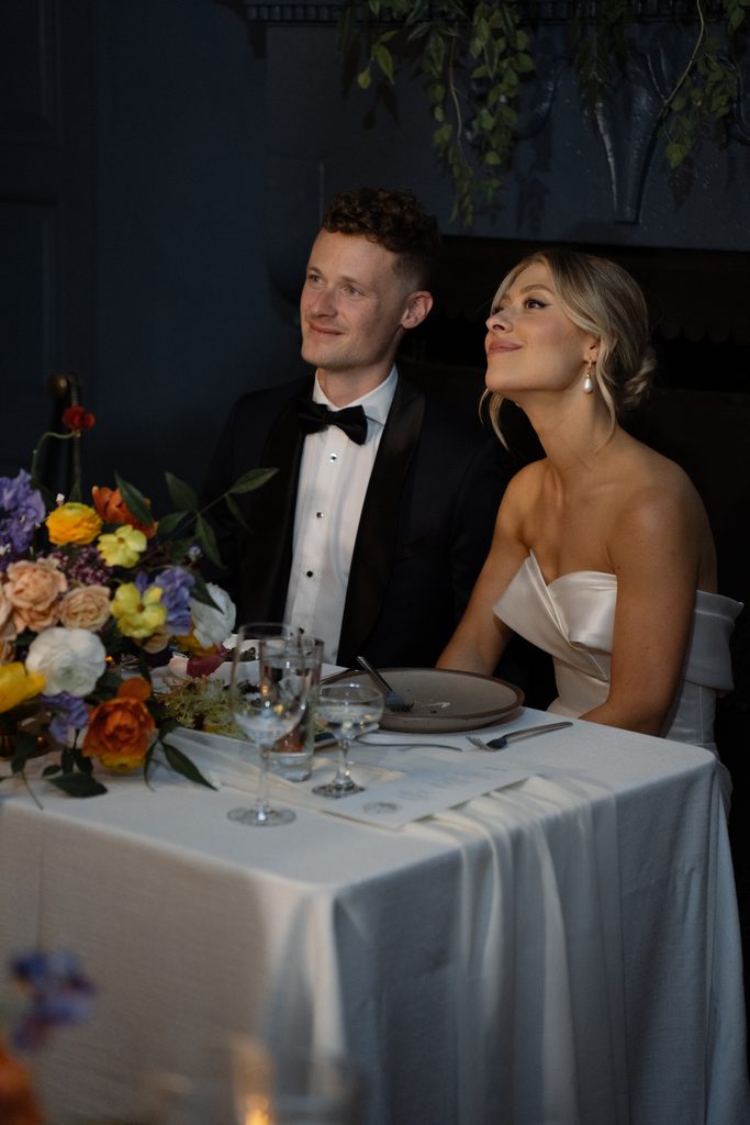 Newlyweds look up lovingly while listening to heartfelt guest speeches at their candlelit sweetheart table.