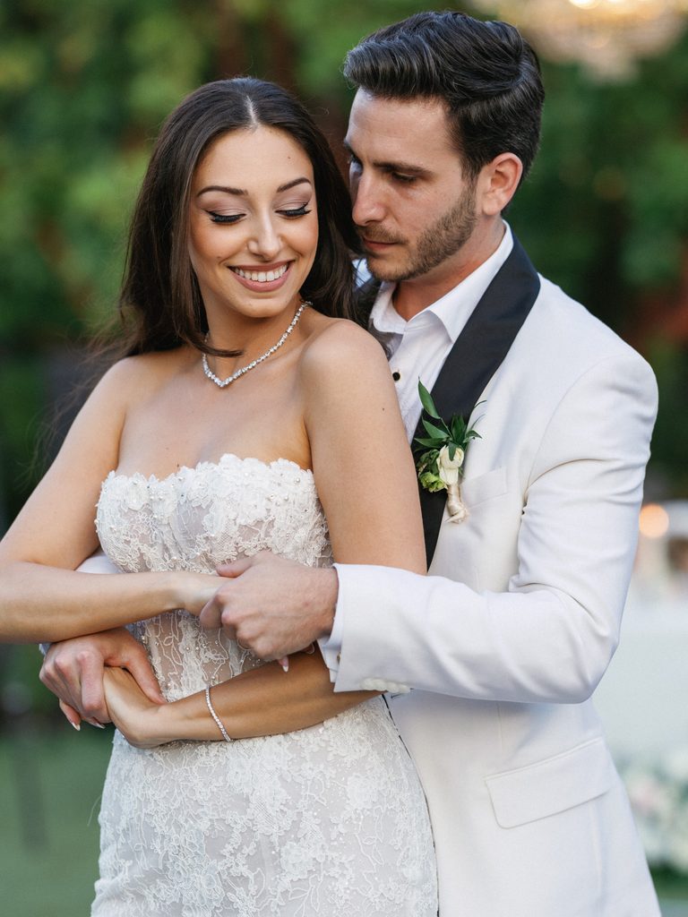 Groom holds the bride from behind during their romantic first dance at a luxury outdoor celebration