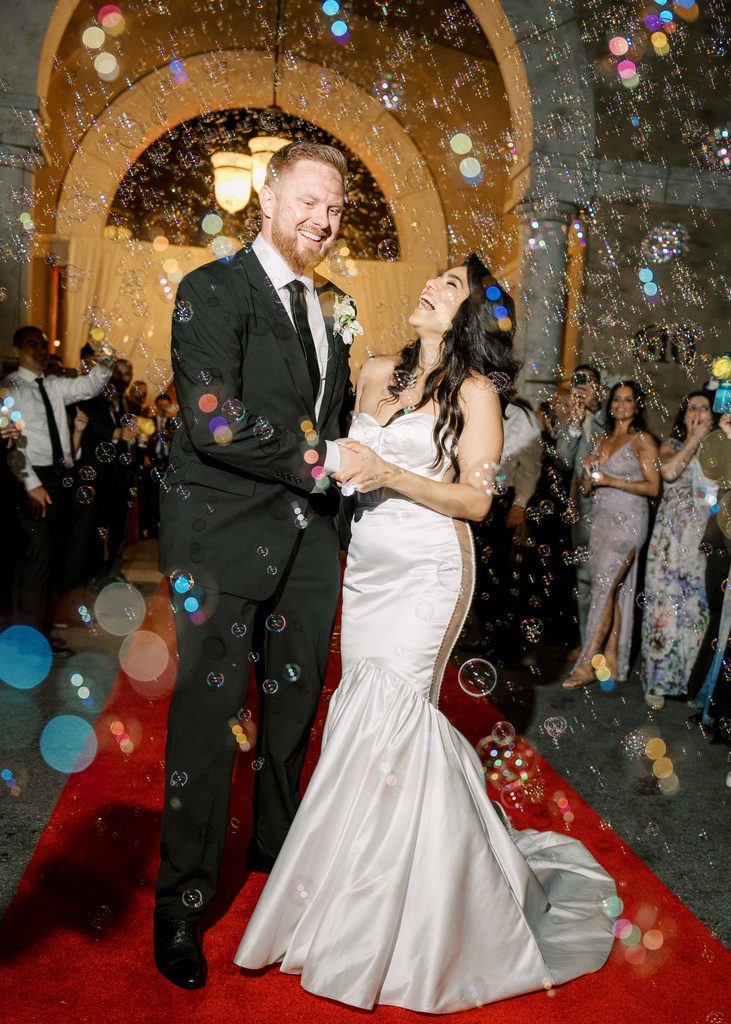 Newlyweds grand exit on a red carpet under dynamic outdoor wedding reception lighting with colorful bubbles.