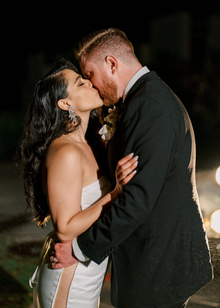 Candid night photo of a bride and groom sharing a romantic kiss, beautifully enhanced by dramatic wedding reception lighting that highlights their silhouettes.