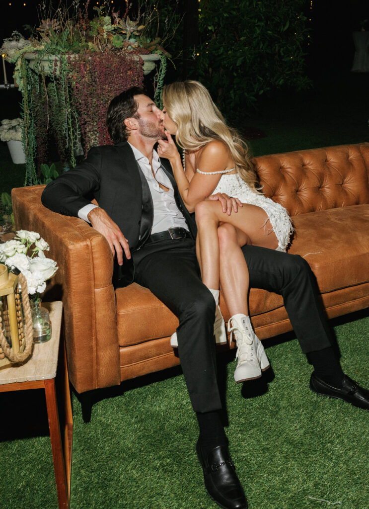 Candid night photo of newlyweds kissing on a leather couch under modern wedding reception lighting.