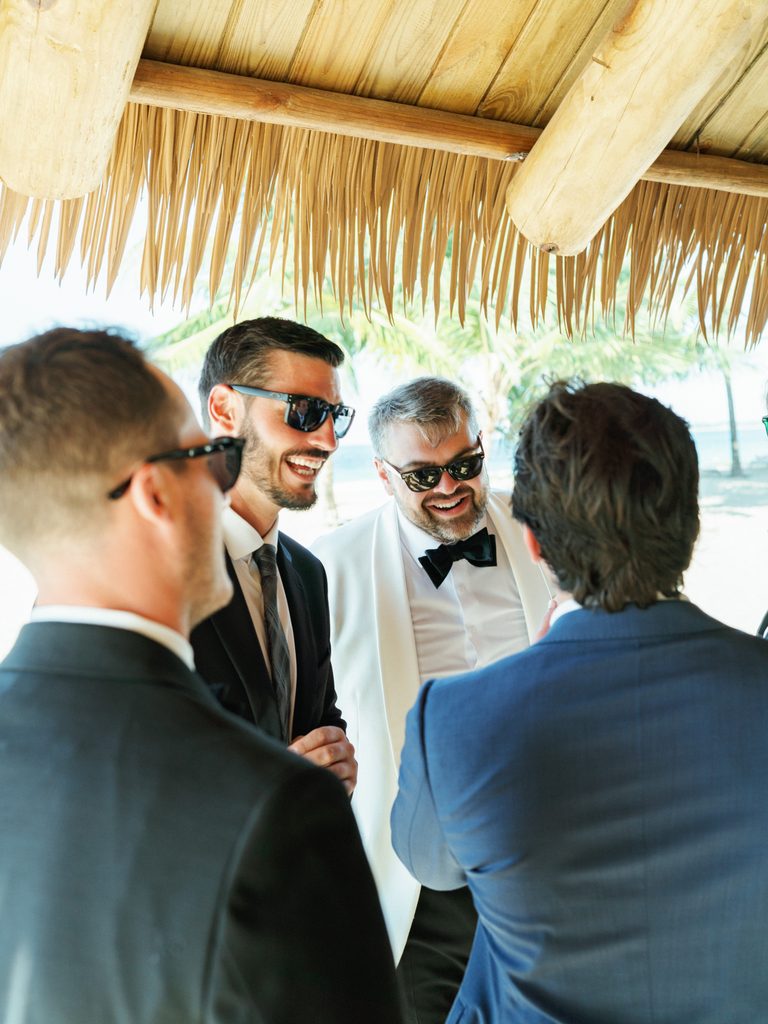 Trendy groomsmen sharing a fun moment under a tropical straw roof at reception