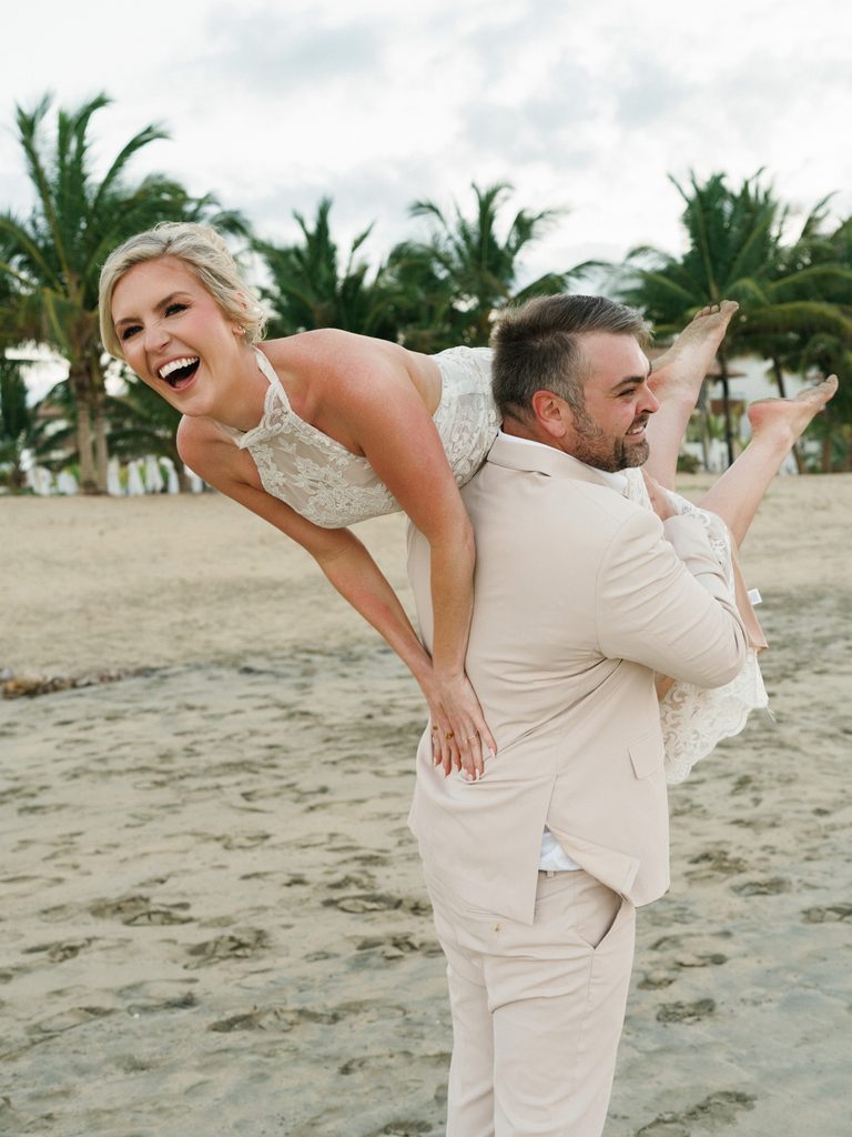 Playful groom lifting his laughing bride on a tropical sand beach with palm trees