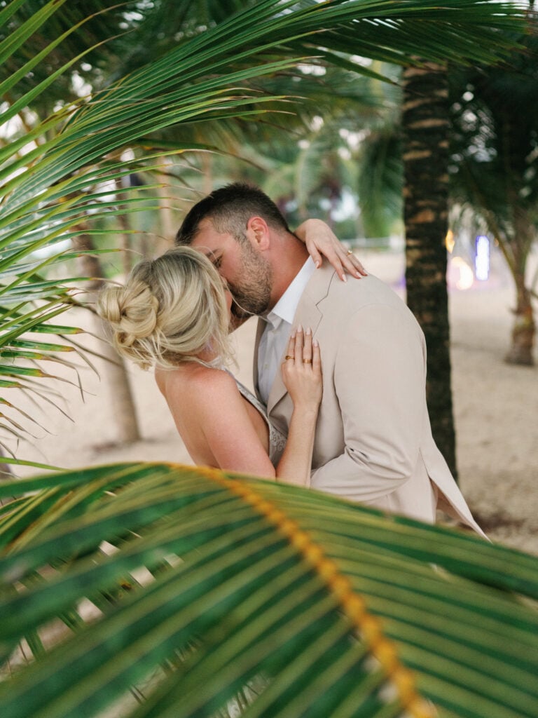 Cinematic bride and groom sharing a beach kiss framed by vibrant exotic palm trees