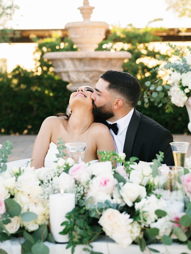Candid wedding reception photos of a joyful bride laughing as the groom kisses her cheek at their table.