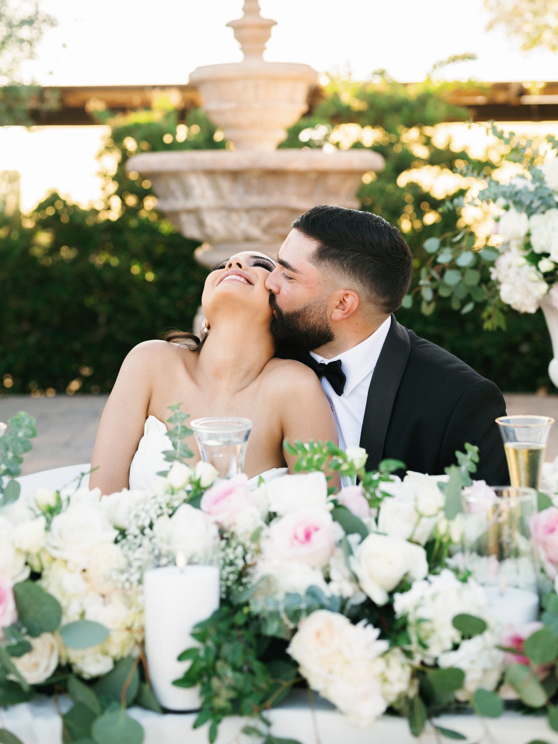 Candid wedding reception photos of a joyful bride laughing as the groom kisses her cheek at their table.