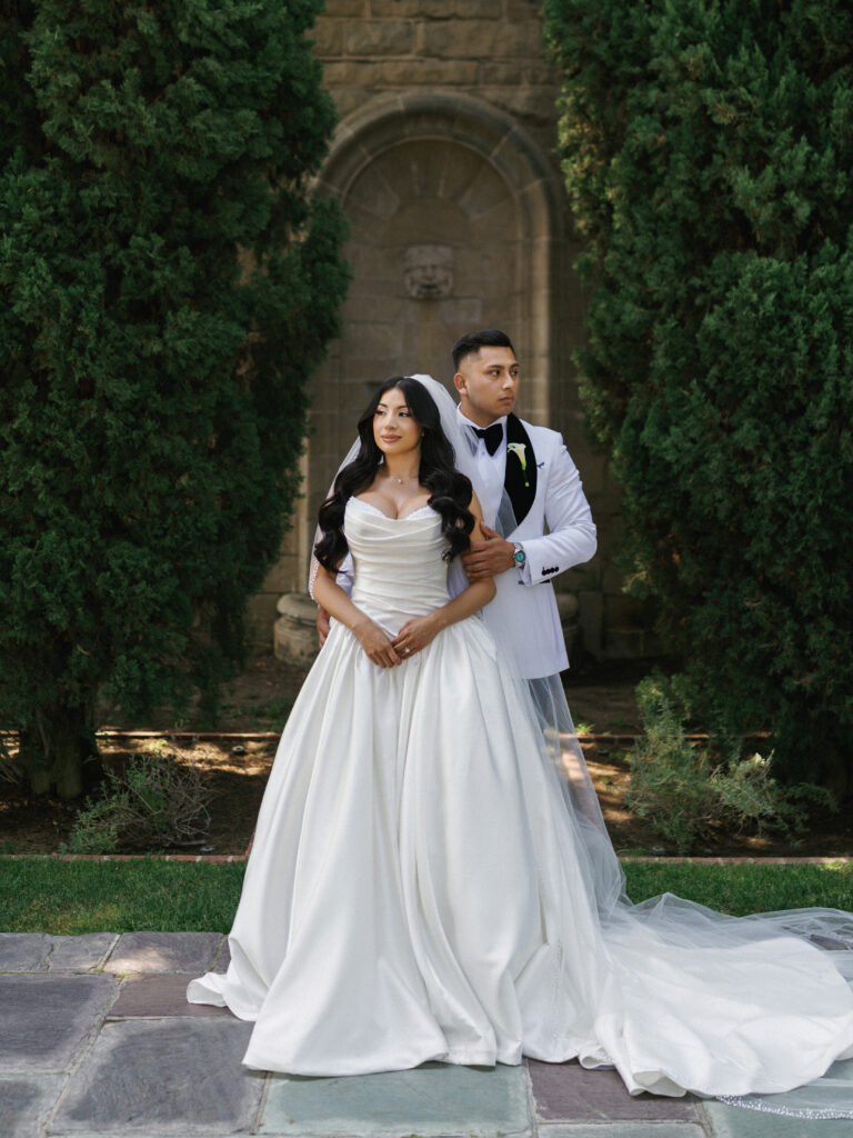Regal newlyweds posing together by a historic stone archway with tall green trees