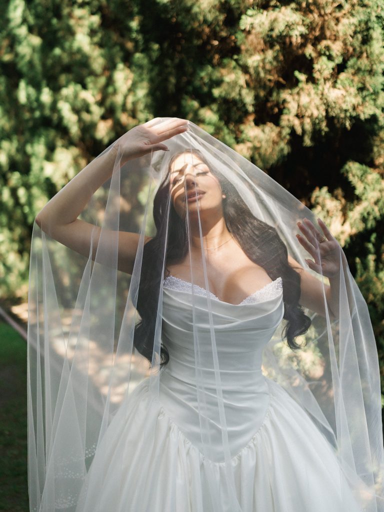 Editorial bridal portrait session in a sunlit garden featuring fine art veil play aesthetics