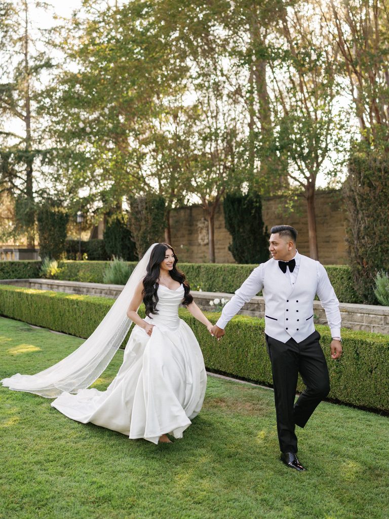 Joyful bride and groom holding hands while running on a manicured estate lawn