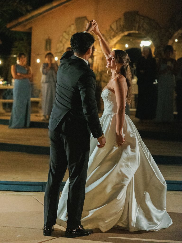Newlyweds dancing outdoors illuminated by romantic wedding reception lighting and string lights.