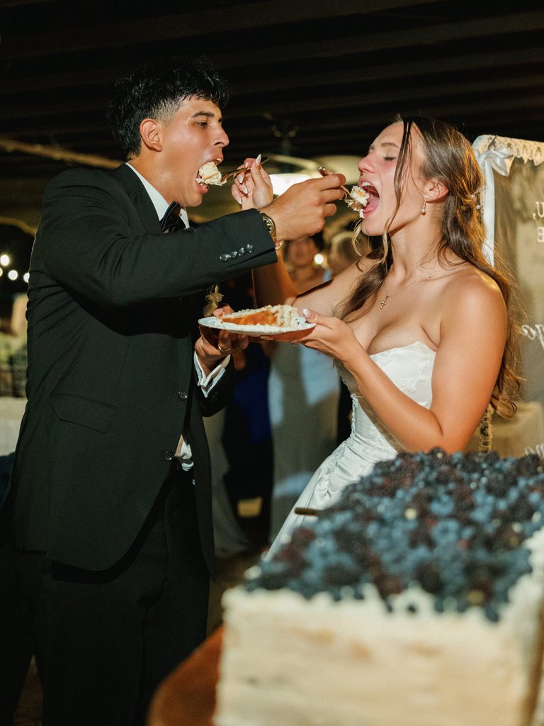 Bride and groom feeding each other cake under crisp wedding reception lighting.