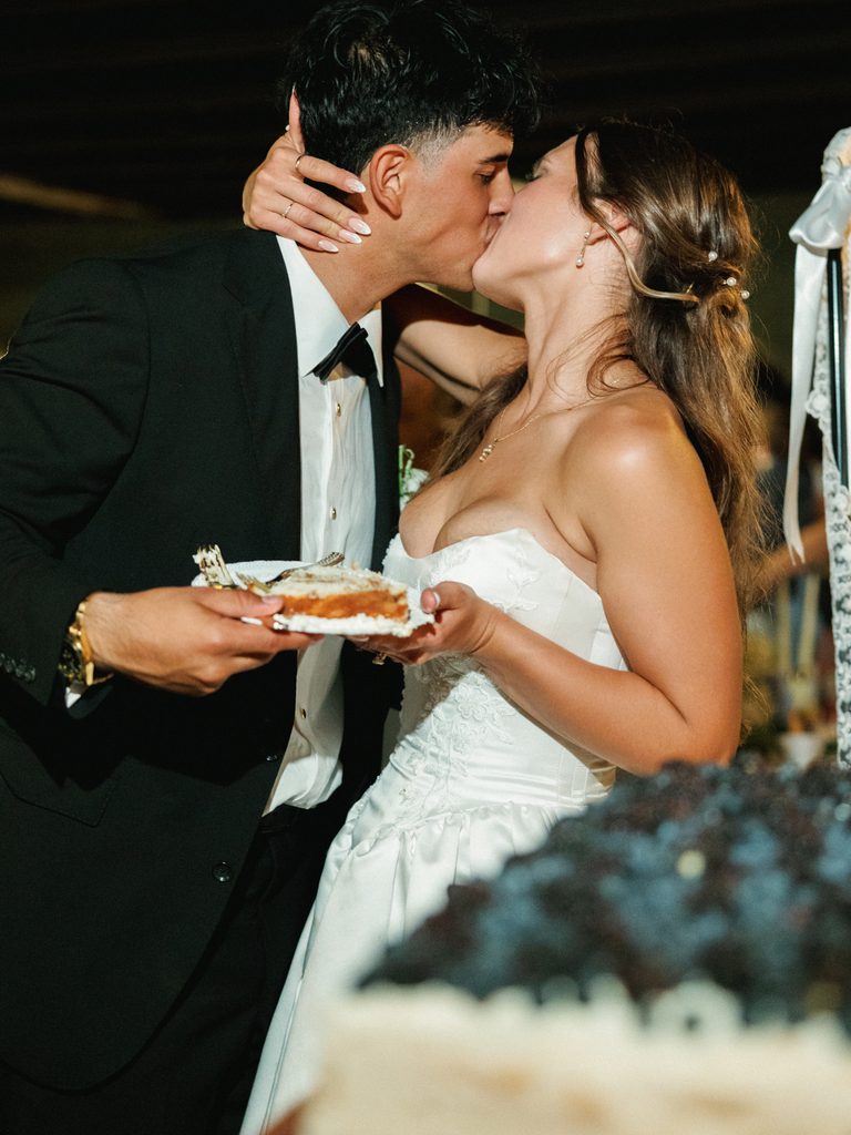 Newlyweds kissing while holding a slice of cake during their wedding reception.