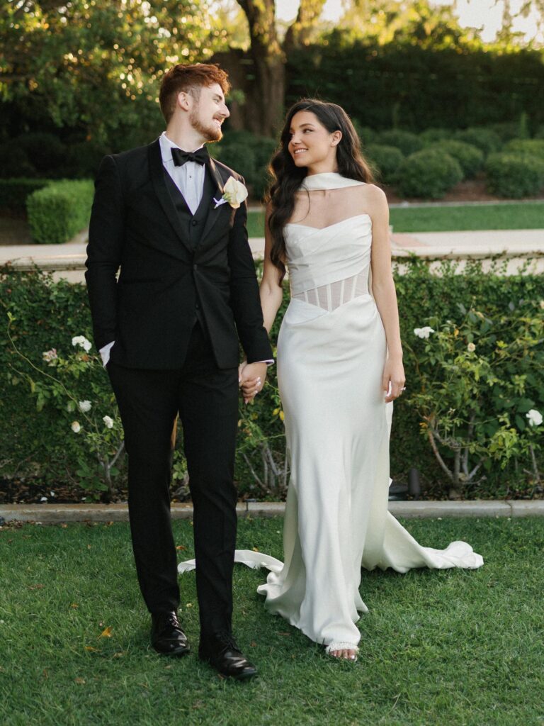 Elegant bride and groom strolling along lush green hedges during their outdoor summer wedding portrait session.
