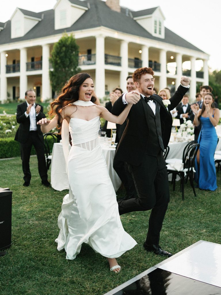 Joyful newlyweds making their grand entrance under soft outdoor wedding reception lighting.