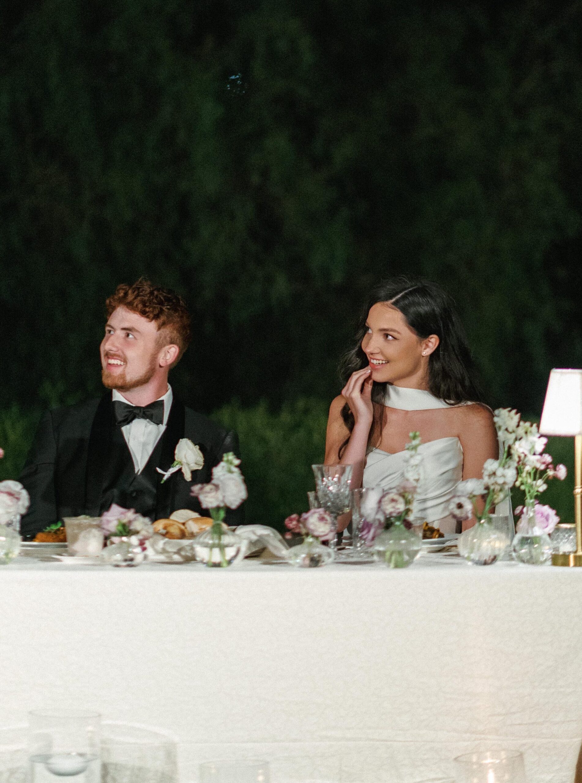 Bride and groom listening to wedding toasts at their sweetheart table under elegant reception lighting.