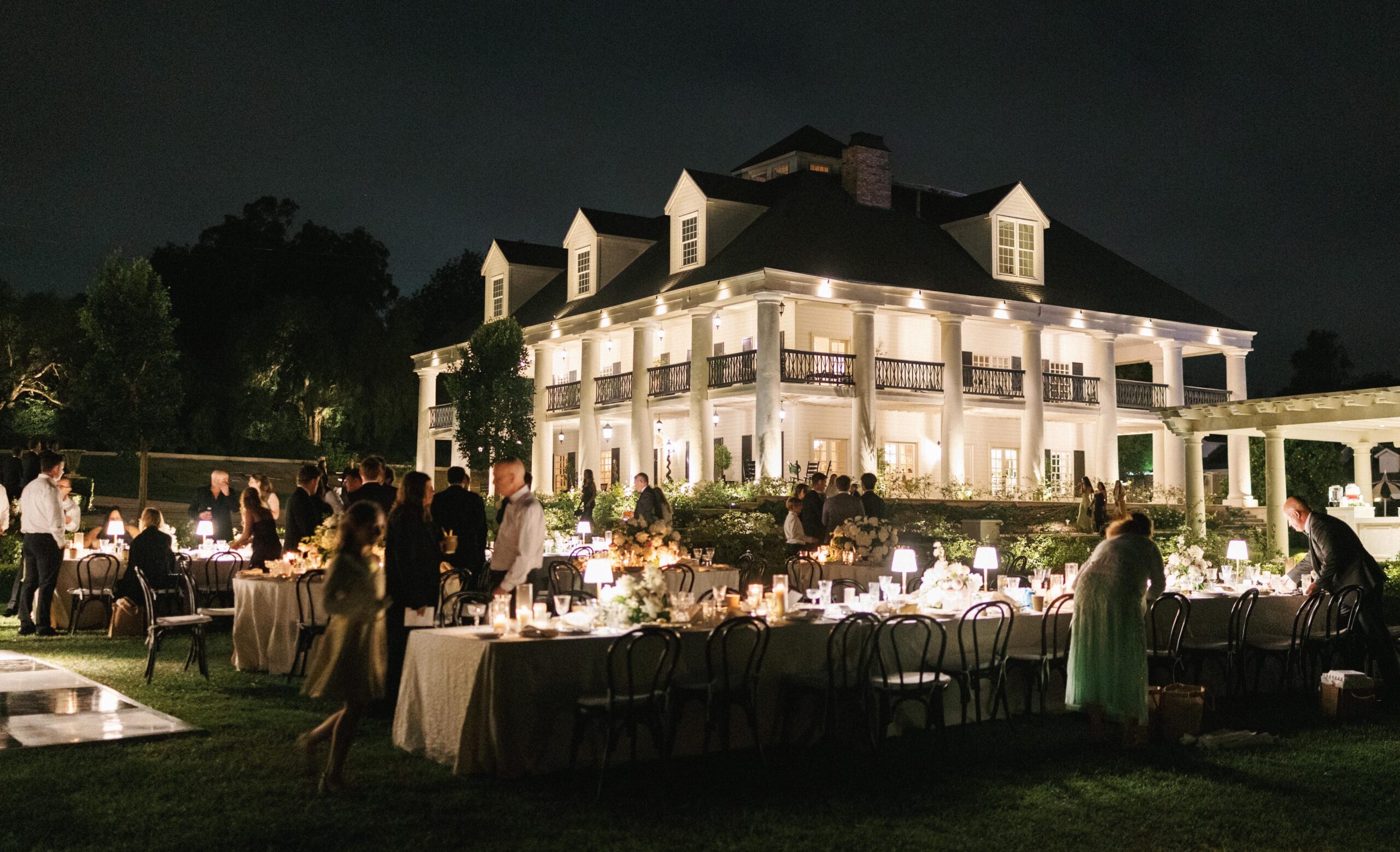 Luxury outdoor wedding reception lighting setup featuring an illuminated estate estate facade and glowing long dinner tables at night.