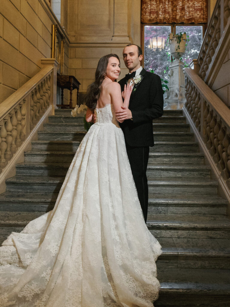 Elegant bride and groom posing on a grand stone staircase inside a historic mansion, demonstrating classic and timeless wedding photography styles.