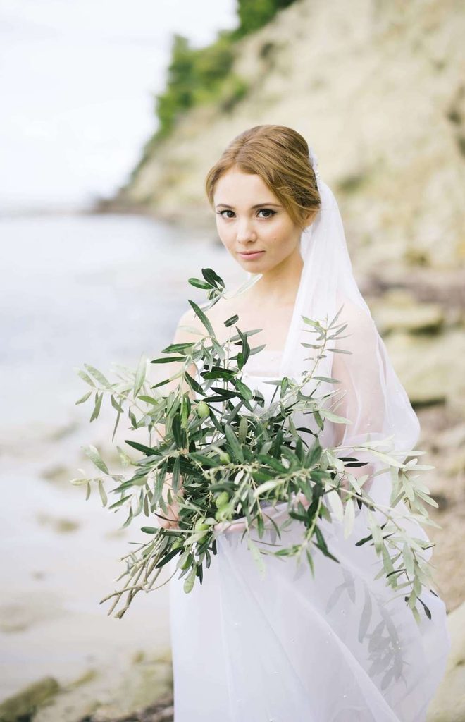Beautiful bride holding an organic olive branch bouquet on a sandy beach