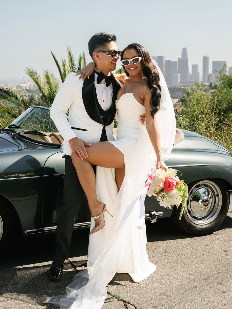 Glamorous bride and groom in sunglasses posing by a vintage Porsche Speedster with the LA skyline behind them, exemplifying cinematic wedding photography styles.