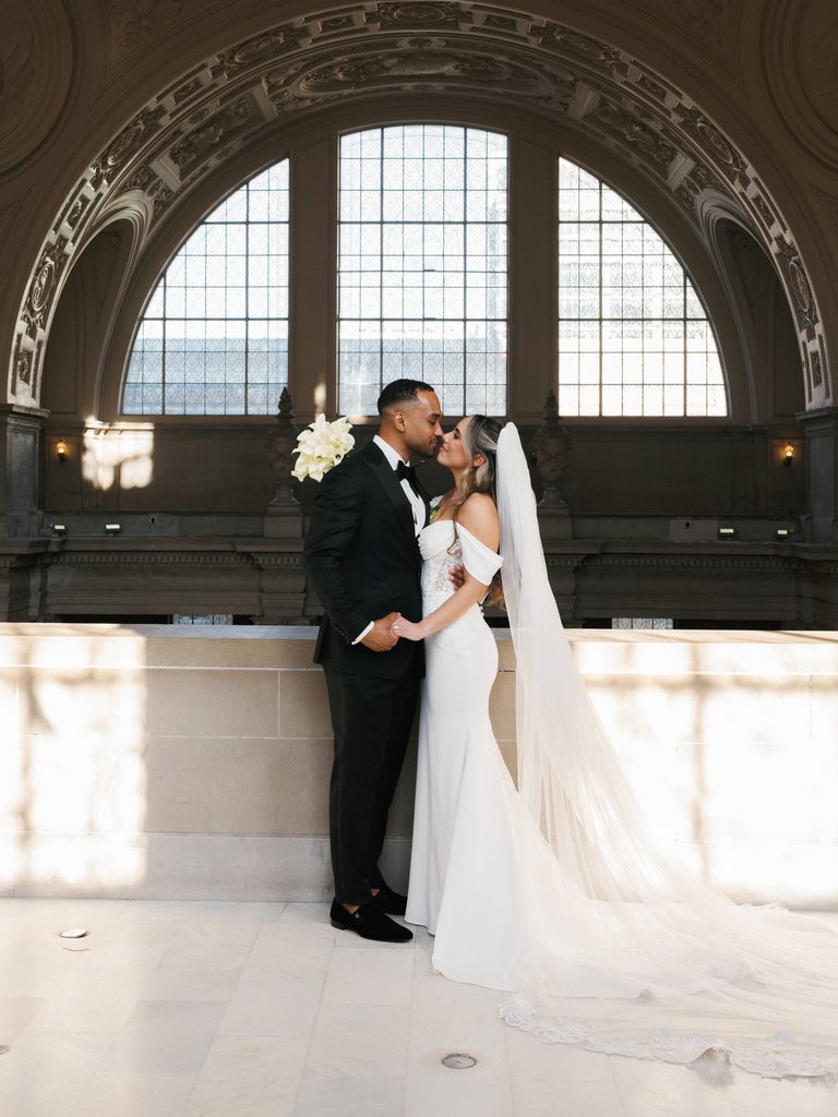 Bride with a long cathedral veil embracing her groom in a sun-drenched historic building, highlighting epic, light-filled wedding photography styles.