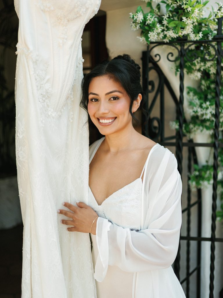 Smiling bride in a white silk robe standing next to her luxury lace gown