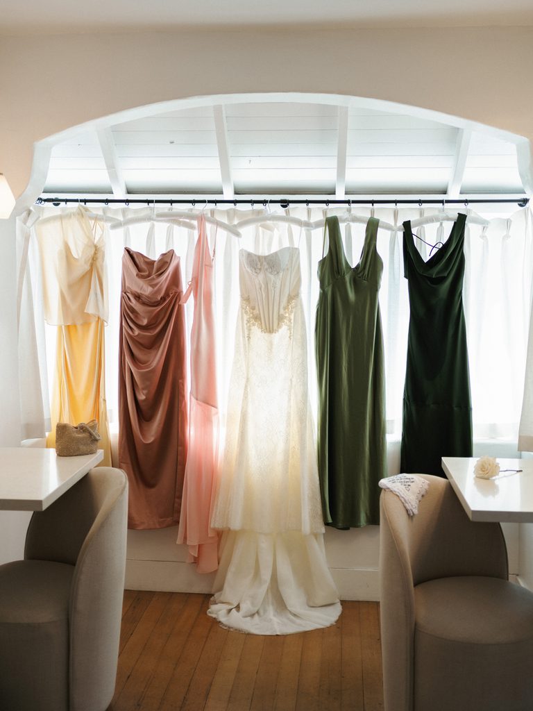 Colorful silk bridesmaid dresses hanging in a row on a white window curtain rod
