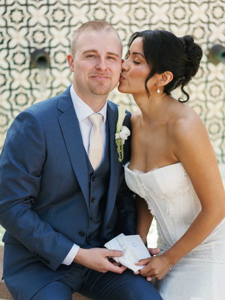 Loving bride kissing her grooms cheek while sitting by a luxury tile fountain