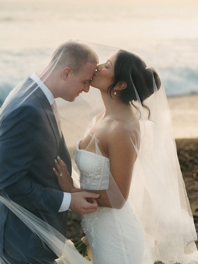 Loving bride kissing her groom under a flowing sheer veil on a sandy beach