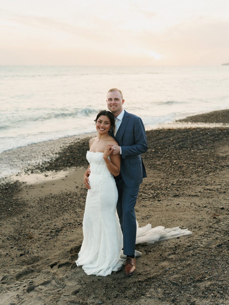 Smiling newlyweds posing on a sandy ocean beach under soft sunset lighting