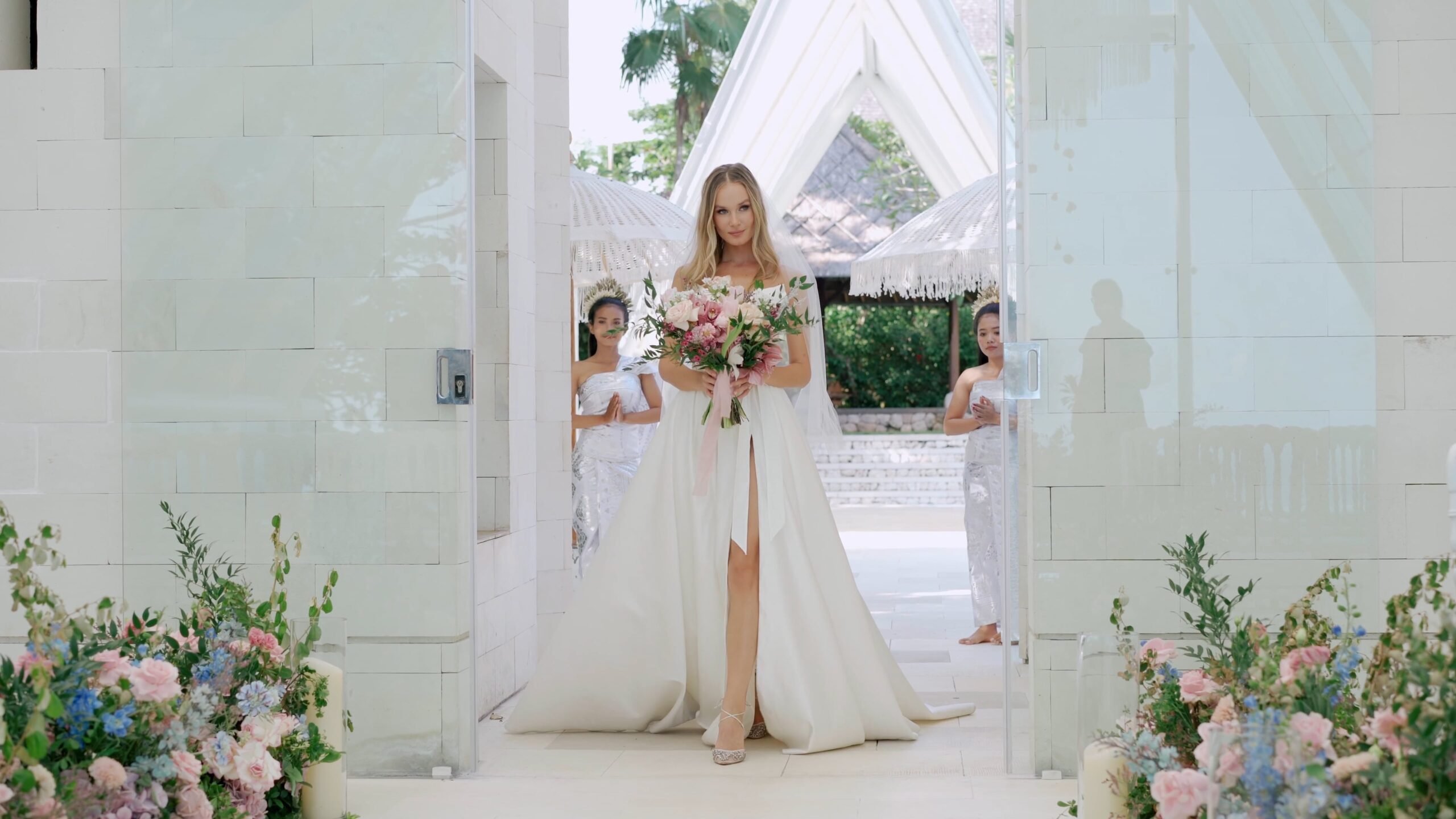 Bride holding flowers at a wide doorway, with two attendants behind her.