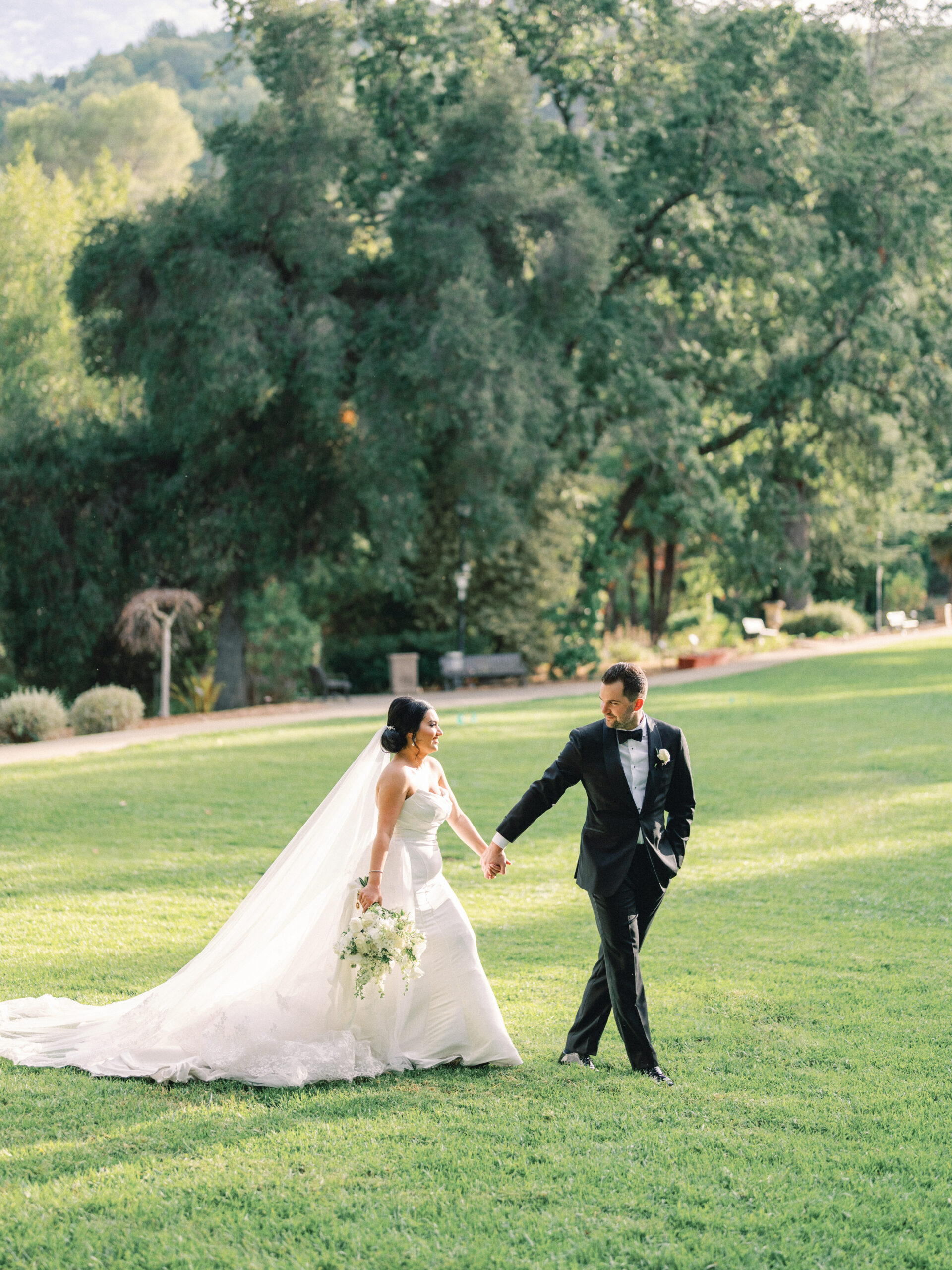 Couple in wedding attire walking across a broad green lawn bordered by trees.