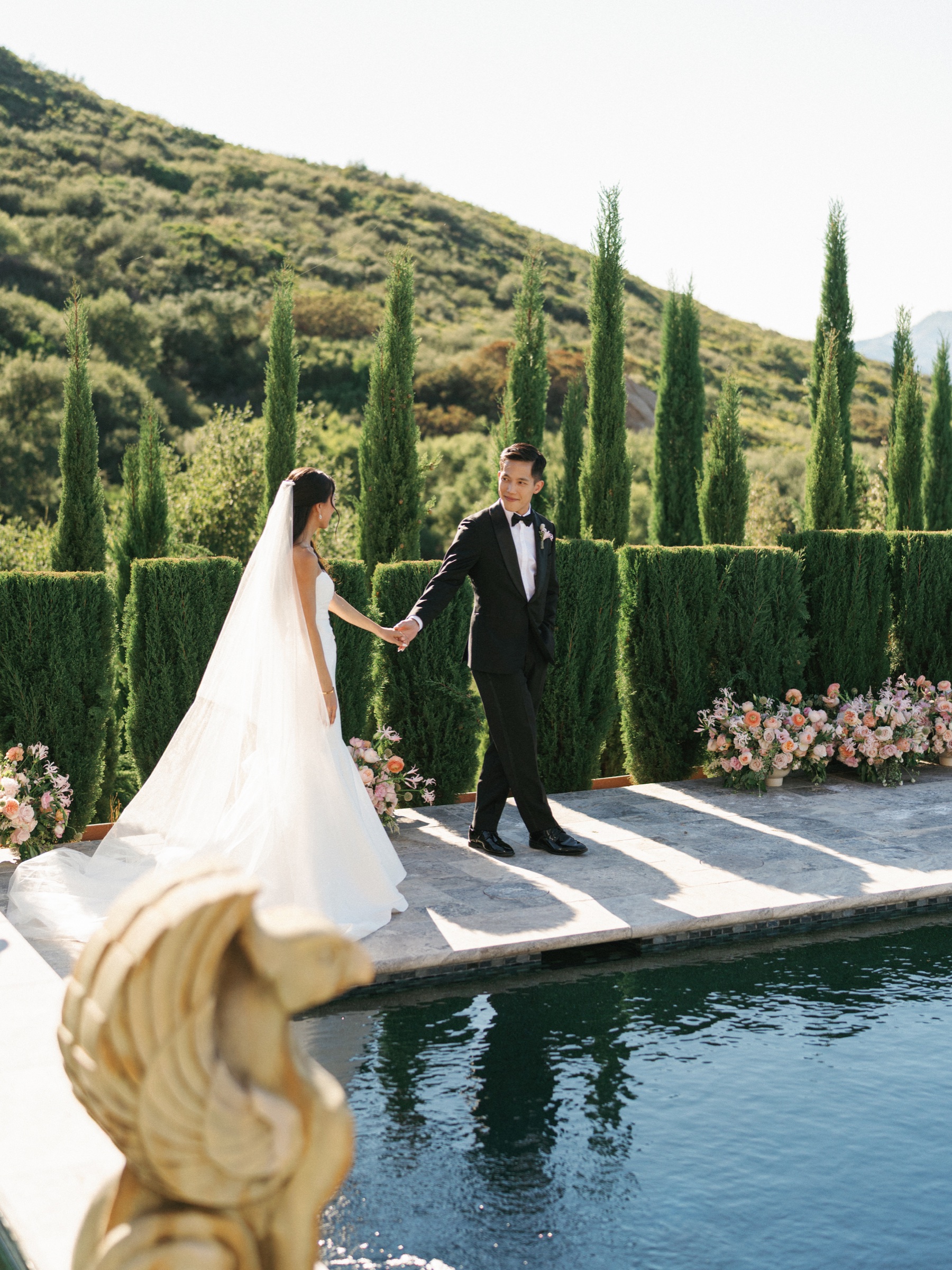 Newlyweds walk beside a pool and cypress-lined hillside