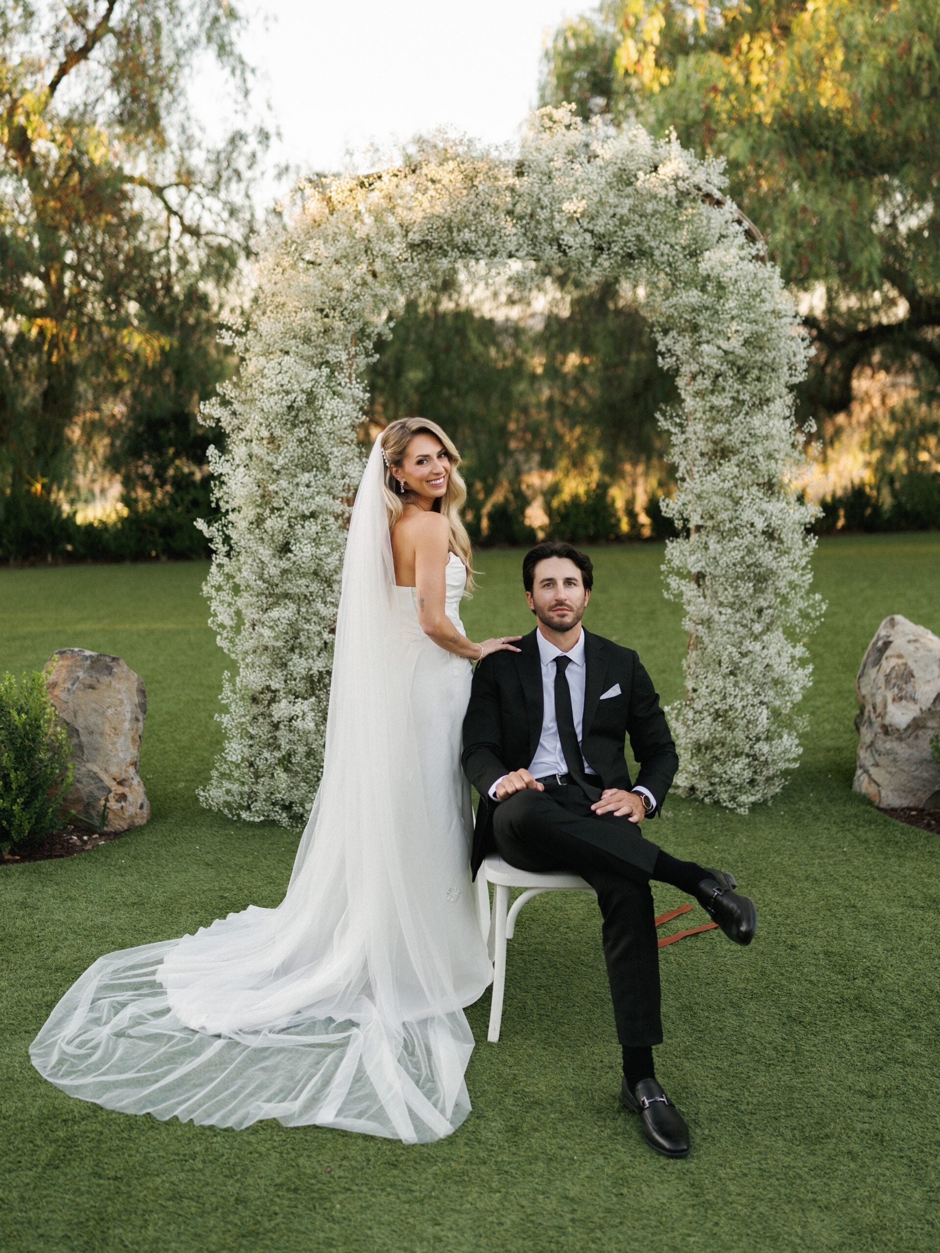 Newlyweds pose beneath a white floral arch on a green lawn.