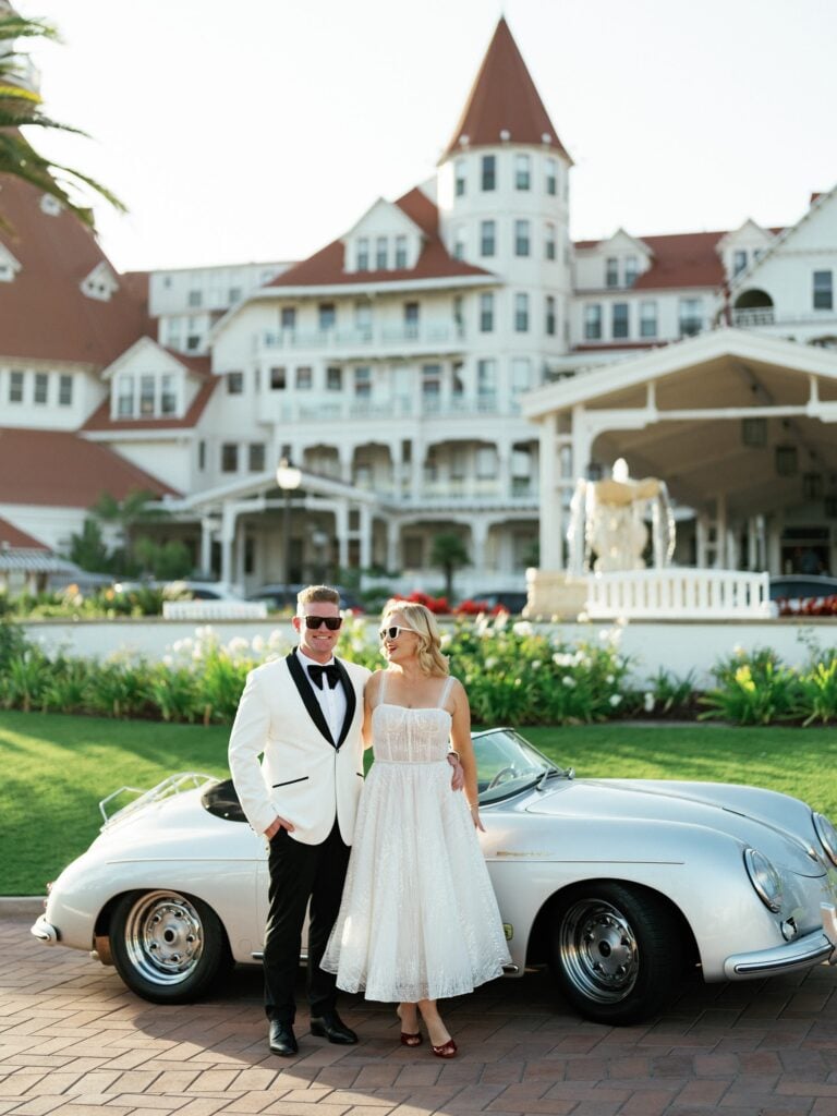 Newlyweds stand beside a vintage car in front of a historic hotel