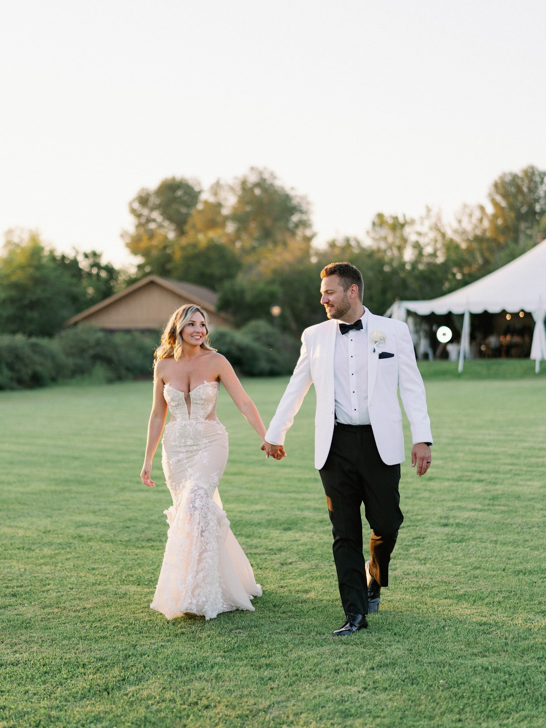 Newlyweds walk across a lawn beside an outdoor reception tent