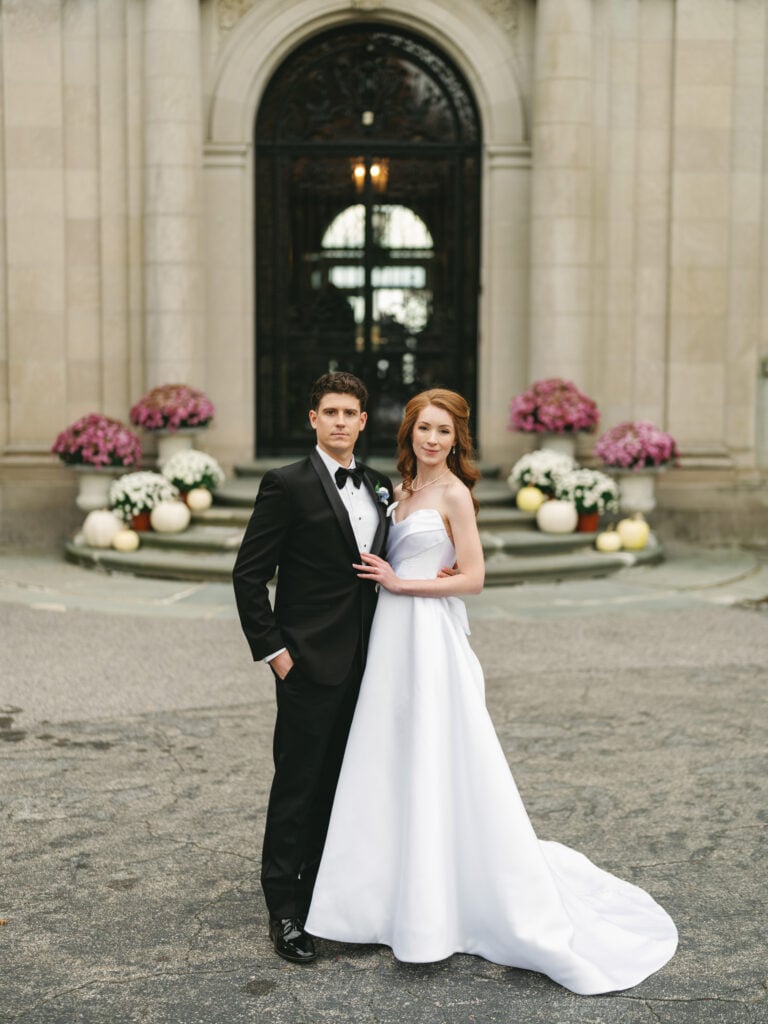 Newlyweds stand together outside a stone building entrance with flower arrangements.