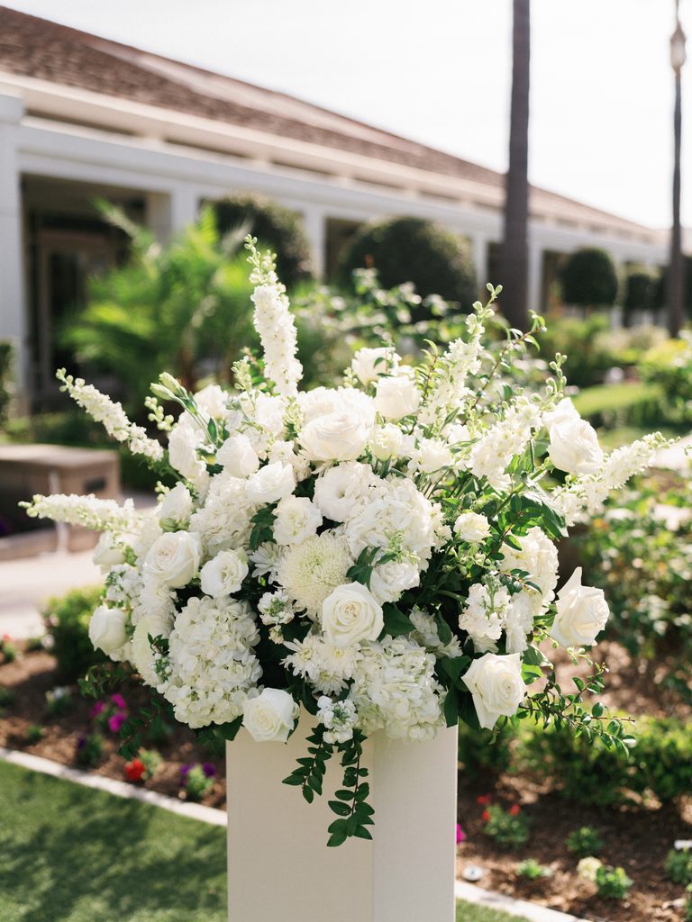 Lush white rose and hydrangea floral arrangement on a sleek modern pedestal decorating a luxury outdoor wedding ceremony altar
