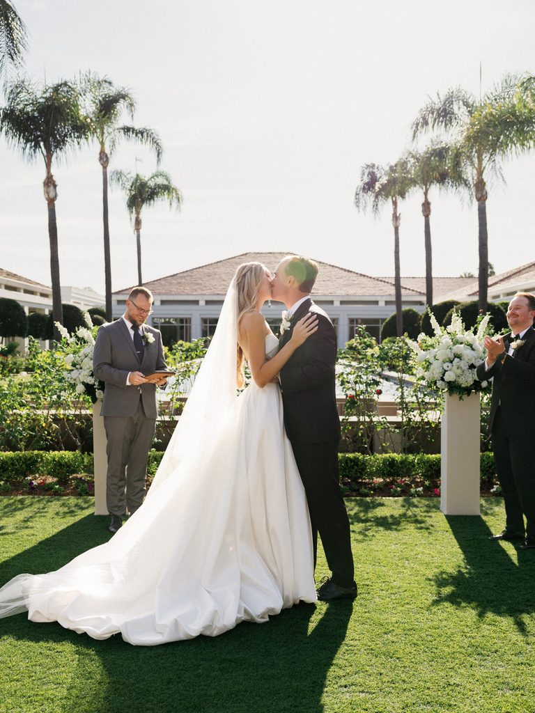 Breathtaking first kiss of a luxury bride and groom during an upscale lawn ceremony with palm trees and white floral pillars