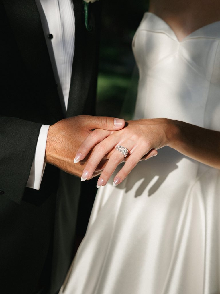 Close up of a groom gently holding his brides hand showcasing a stunning luxury diamond engagement ring set against a white satin bridal gown
